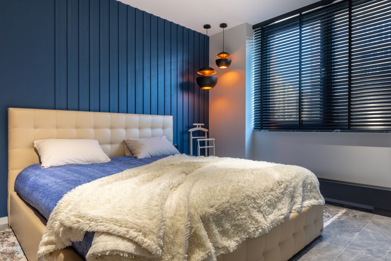 Modern bedroom interior with blanket and pillows on bed against lamps and jalousie in house